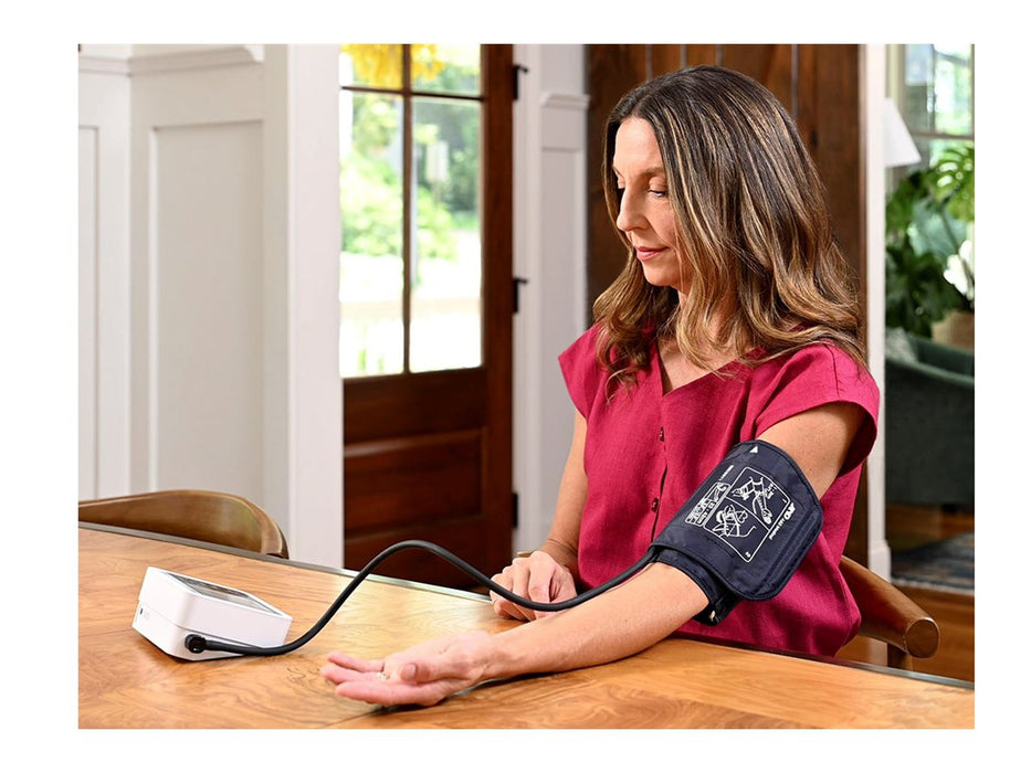 Woman using an upper arm blood pressure monitor with cuff at a dining table in a bright home setting