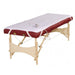 Angled view of a portable wooden massage table smoothly fitted with the white fleece Deluxe Massage Table Warmer Pad, secured with elastic corner straps to eliminate cold surface shock for clients.